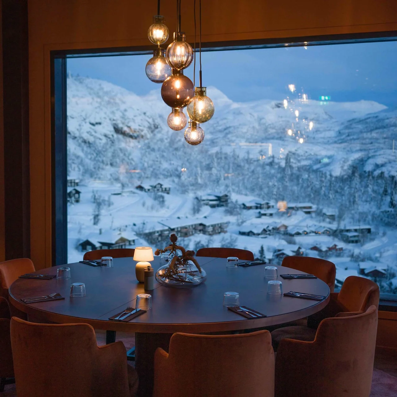 A round dining table, set with glasses and cutlery, under warm hanging lights. Beyond a large window, snow-covered mountains and chalets create a serene winter landscape.