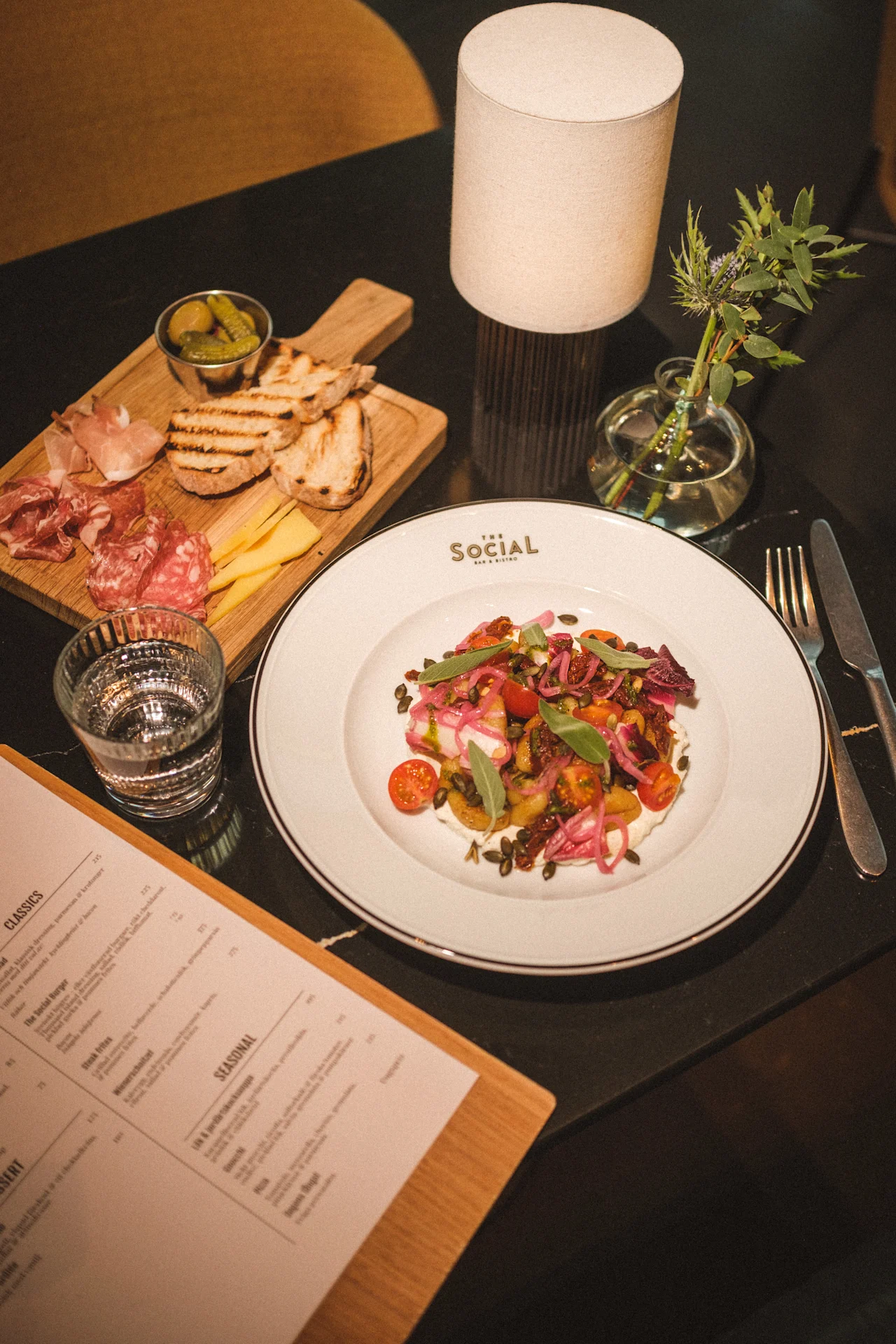 Et lækkert måltid serveret på et bord, med en farverig salat, charcuteri, ost, brød og en menu for THE SOCIAL BAR AND BISTRO.