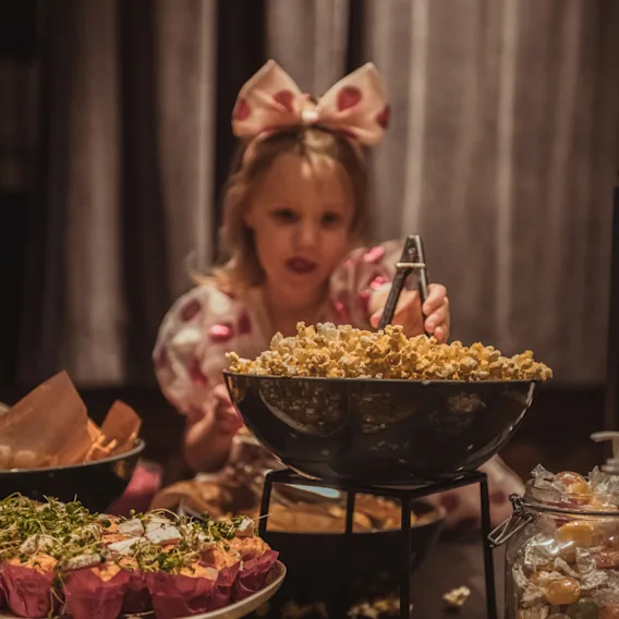 En liten jente med rosa sløyfe i håret forsyner seg med popcorn fra en stor skål på en buffet fylt med muffins, godteri og pizza.