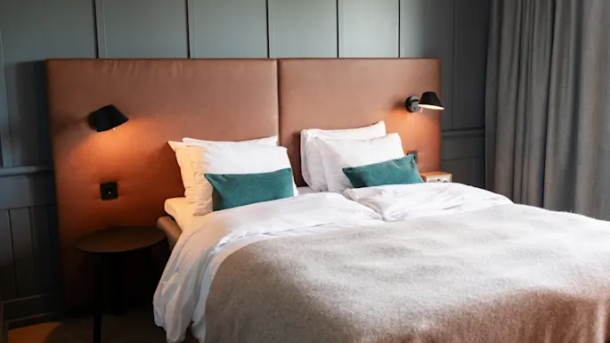 Bed rests under brown headboard with reading lamps, adorned with white sheets and green cushions. Nightstands flank the bed in a softly lit bedroom with gray paneled walls.