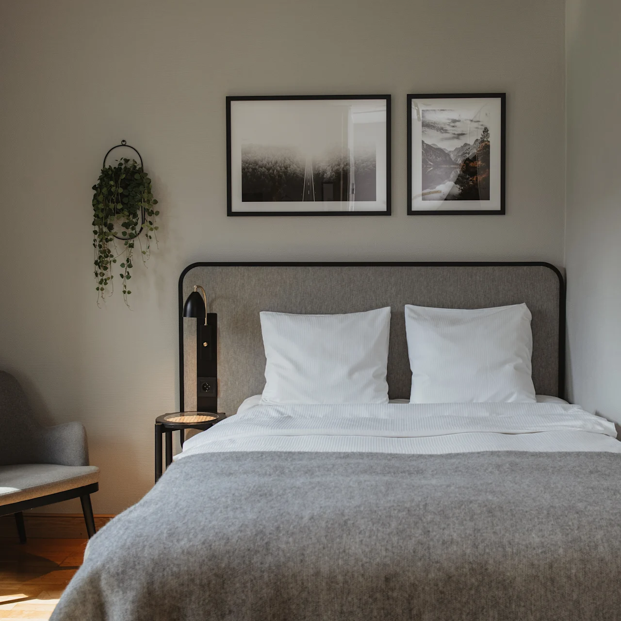 A neatly made bed with white pillows and a gray blanket stands in a small room. Framed pictures hang above, and a hanging plant is on the wall beside a cozy chair.