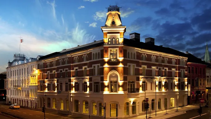 A multi-story building with arched windows is illuminated at night. It stands on a street corner, surrounded by similar architecture, under a twilight blue sky.