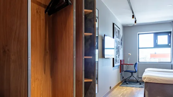 A wooden wardrobe with a hinged door open, revealing clothes hangers, in a modern hotel room with a bed and desk.