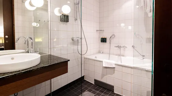 Modern bathroom features a bathtub with a handheld shower, surrounded by white tiles. Double sinks with mirrors on a granite countertop are on the side, under warm globe lights.