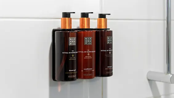 Three dark amber bottles with copper dispensers are aligned on a shelf, labeled as shower gel, conditioner, and shampoo, set against a tiled bathroom wall.