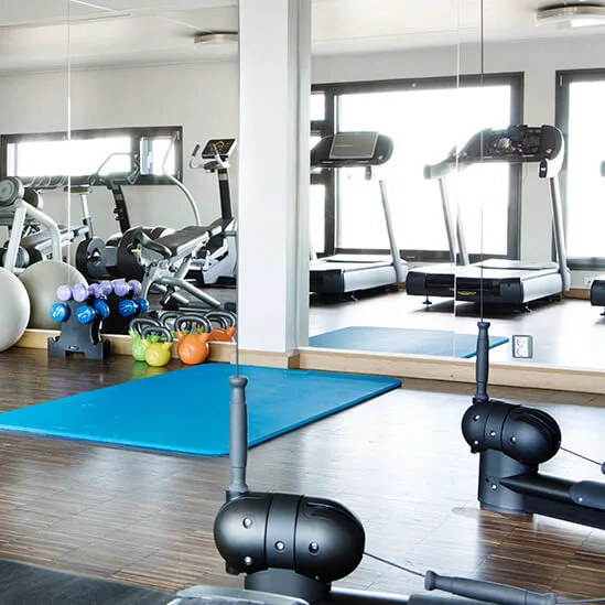 Gym equipment includes treadmills, free weights, kettlebells, and exercise balls on dark wood flooring, with mirrors reflecting the space.