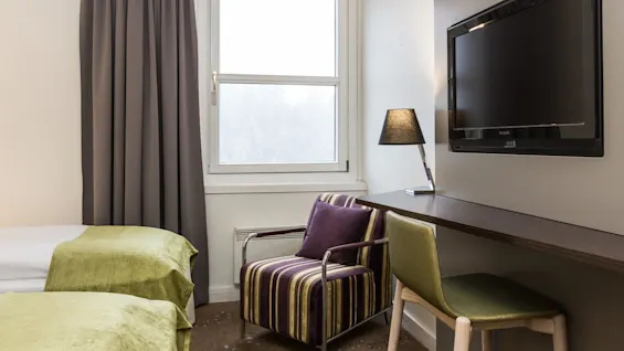 A wall-mounted TV is opposite a striped armchair near a window. A desk with a lamp and chair are beside two beds with green covers.