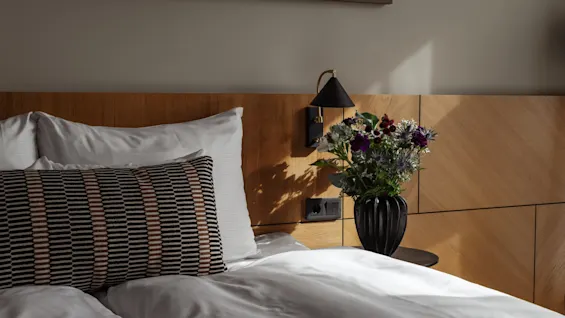 Et koselig hotellrom med seng, mønstret pute, nattbordslampe og en vase med blomster.