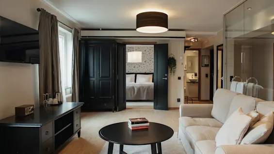 A comfortable hotel suite featuring a living space with a sofa and a bedroom beyond open doors, offering a relaxing stay.