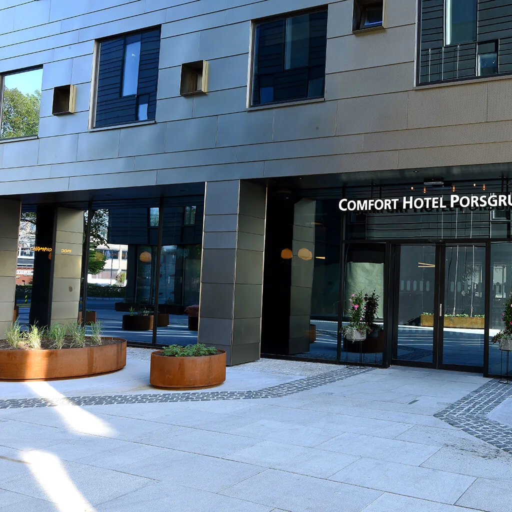 Indgang med glasdøre, der viser "COMFORT HOTEL PORSGRUNN" i bymiljø, omgivet af store plantekasser med grønne planter og moderne arkitektur.