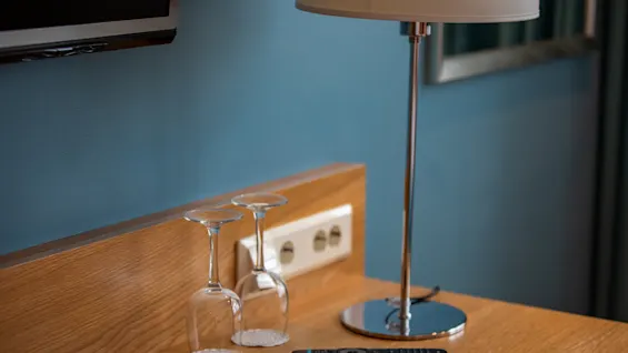 En bordlampe står på en treflate ved siden av to opp-ned-vendte glass og en fjernkontroll, med en TV montert på en blå vegg.