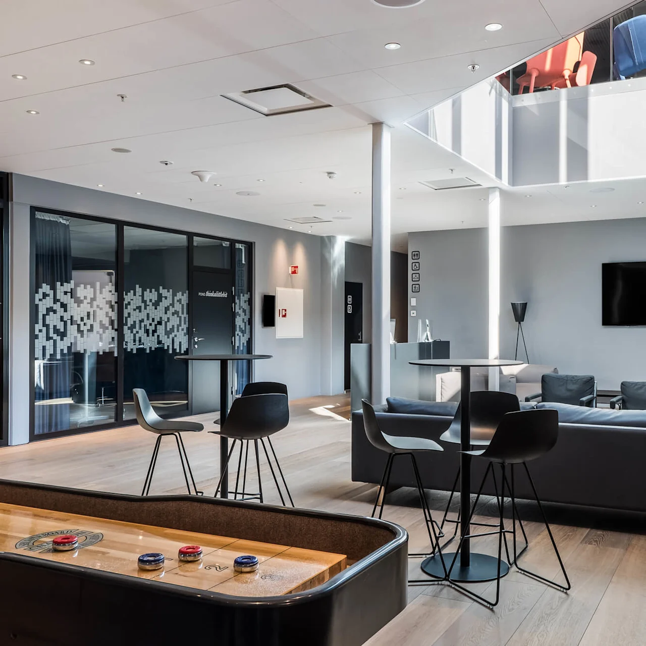 A modern lounge area contains black chairs and a shuffleboard table. Glass walls with abstract designs and a black TV are present. Light floods from above, revealing upper-level chairs.