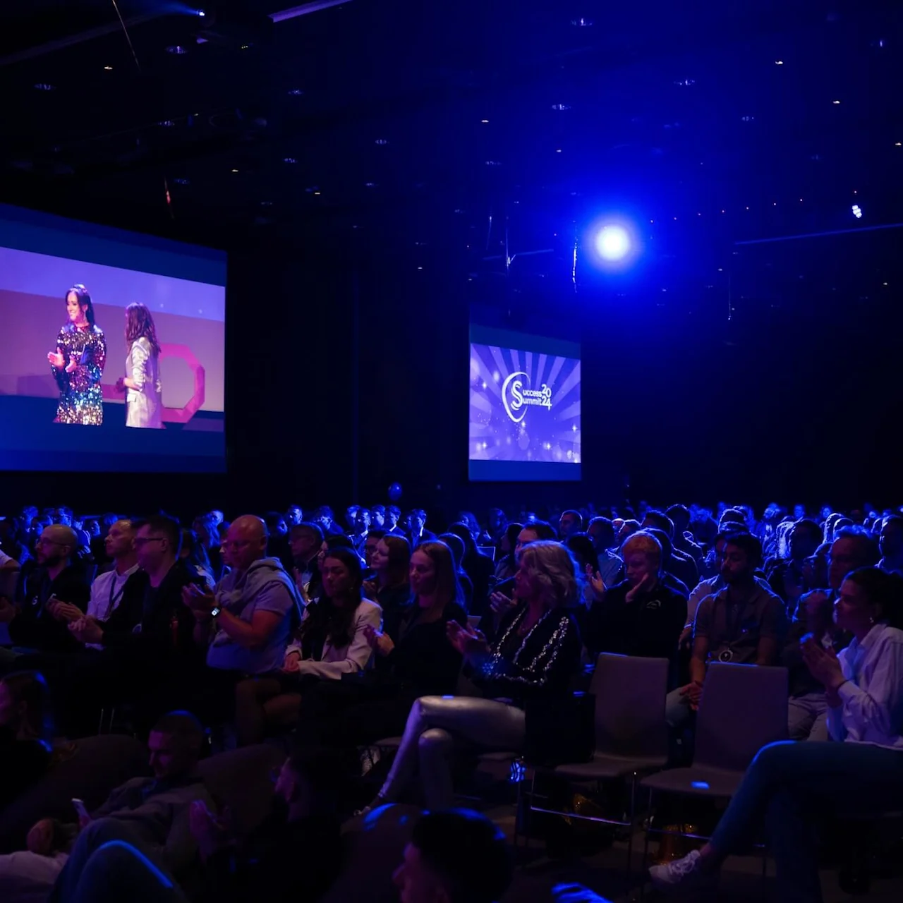 A seated audience claps in a dark auditorium with blue lighting. Two large screens display presenters. Visible text: "Stockholm 20 Summit 24".
