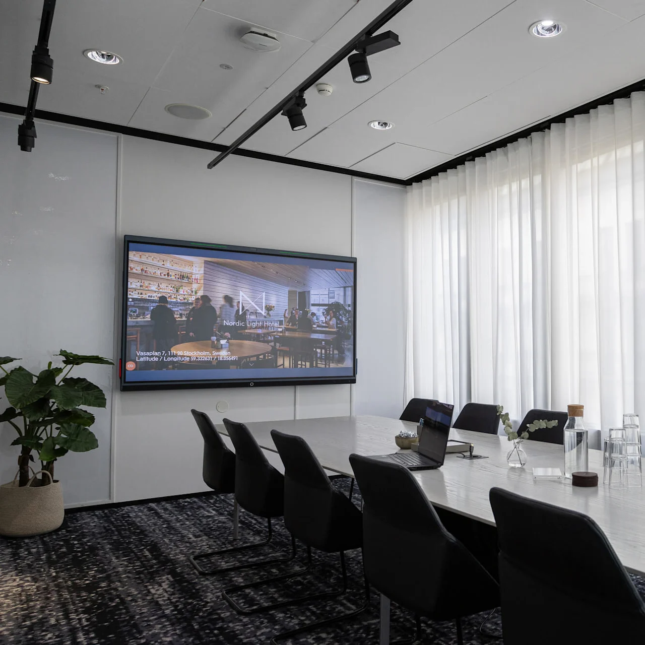 Meeting room features a long table with chairs, a laptop open, glass bottles, cups, a large screen displaying "Nordic Light Hotel" text. Brightly lit by natural light through curtains.