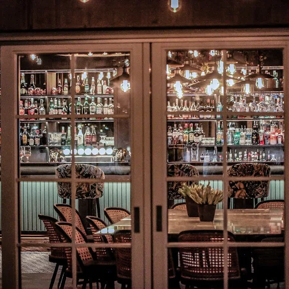 En godt fyldt bar med glashylder fyldt med flasker og glas, bag et bord med fletstole, varmt oplyst i en elegant kontekst.