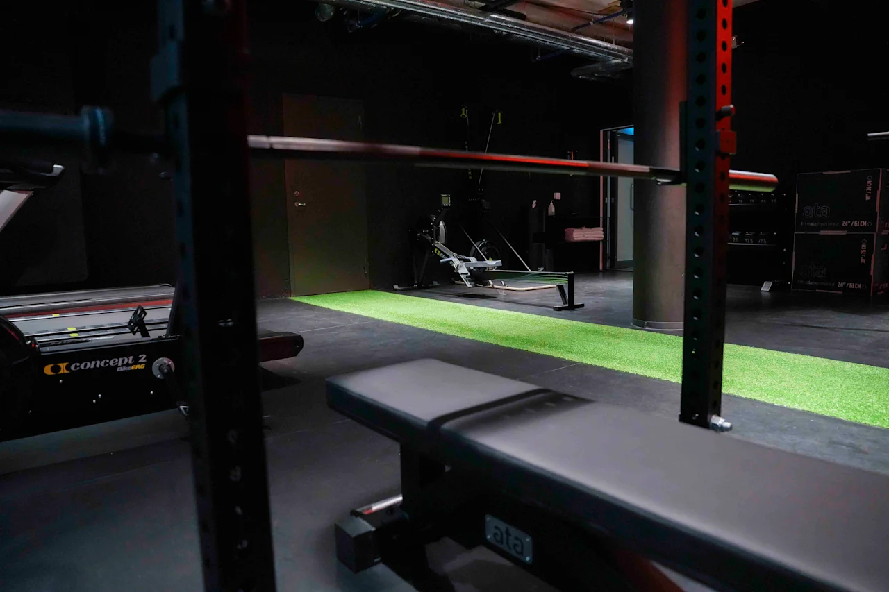 En vægtbænk står tom under en vægtstang i et dæmpet oplyst fitnesscenter, omgivet af træningsudstyr og et stykke grønt kunstgræs.
