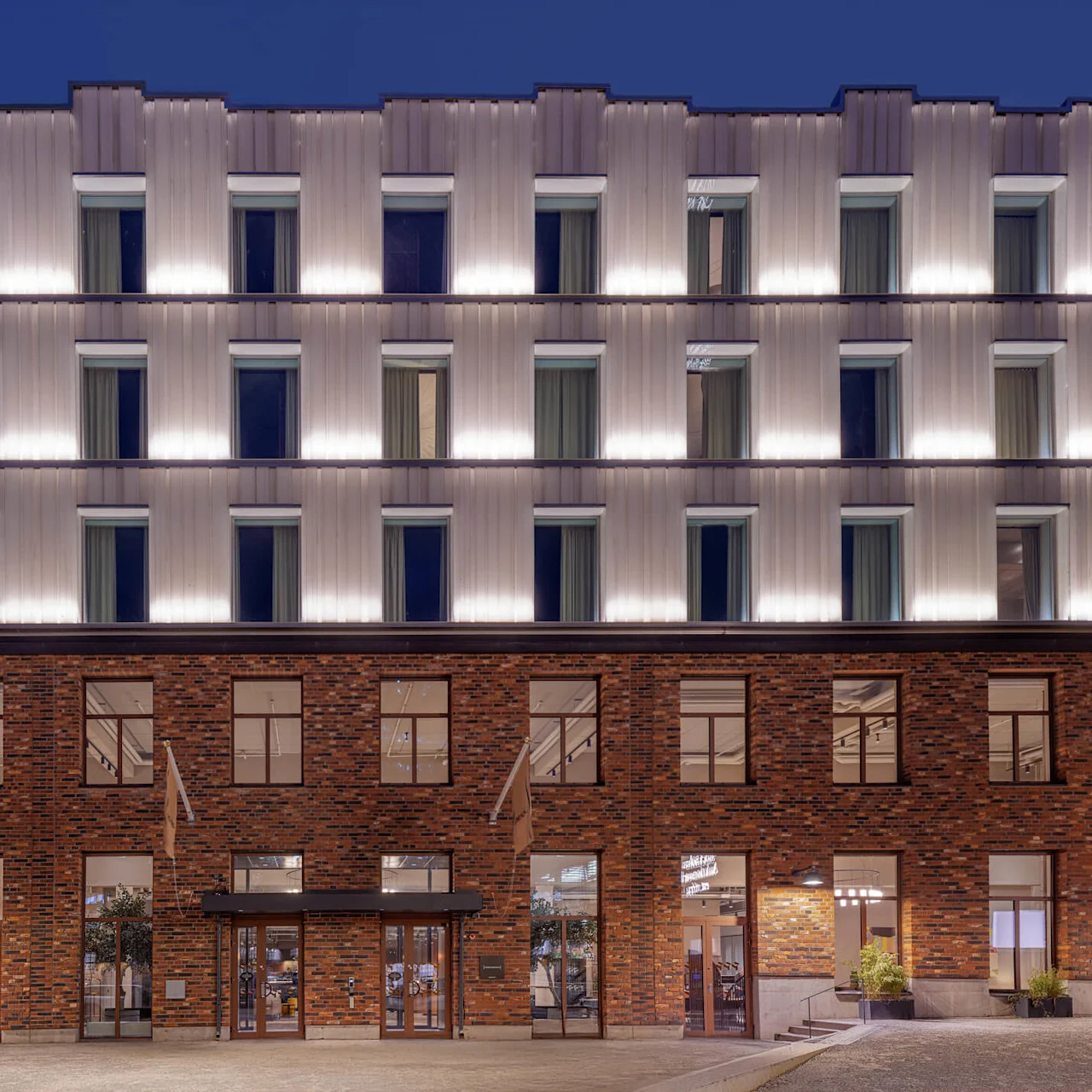 A modern building facade features illuminated windows on upper metal-clad levels and a brick lower level, set against a clear evening sky.