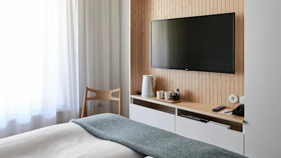 A wall-mounted TV above a wooden shelf with a kettle, cups, and remote in a minimalist room. A bed with a blue blanket and curtains add softness to the space.