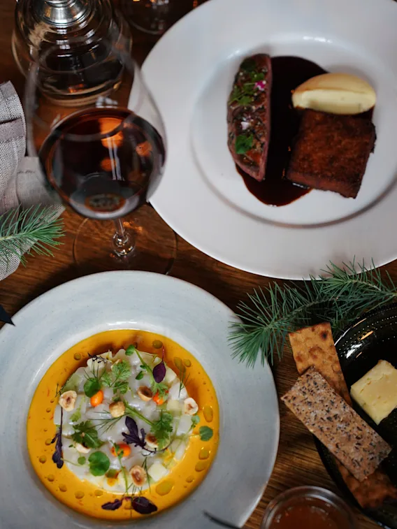 Plates with gourmet dishes: one features sliced meat, mashed potatoes, and sauce, another with colorful garnished salad; accompanied by a glass of red wine and assorted appetizers on a wooden table.