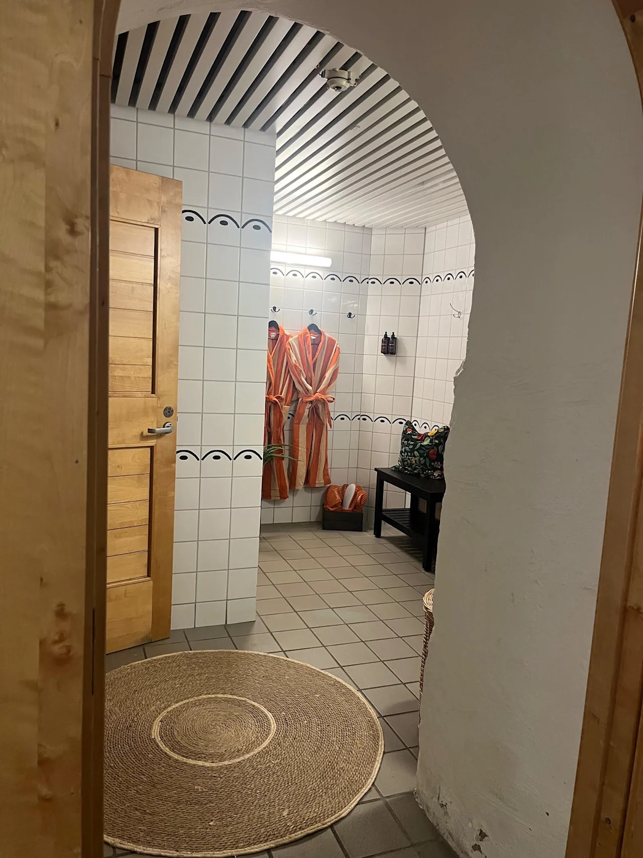 Et flislagt velværeområde med to badekåper hengende, en benk med en pute og en rund matte på gulvet, som inviterer til avslapning.