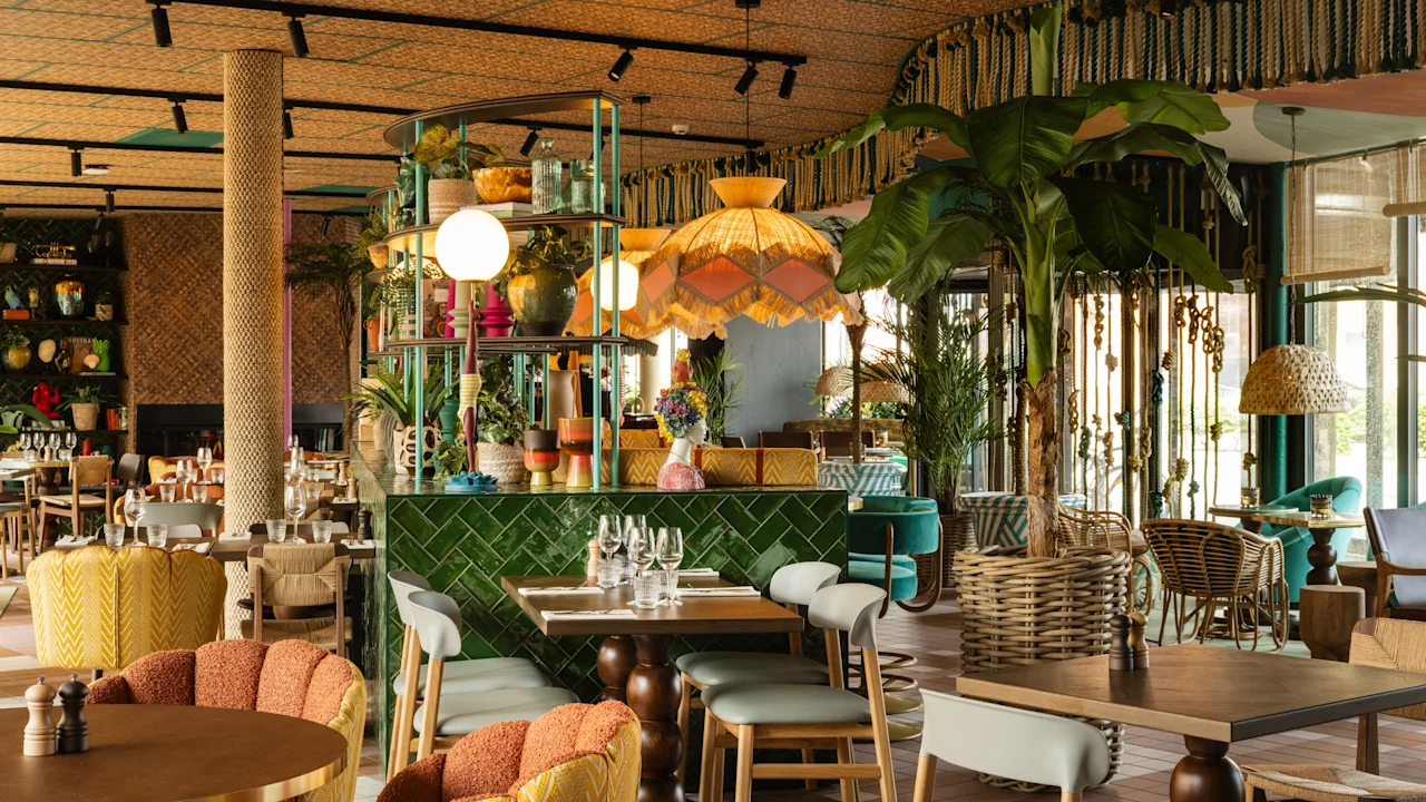 En restaurantindretning har farverig, eklektisk interiør med frodige planter, træmøbler og blød belysning. Bordene er sat med glas og bestik, hvilket skaber en indbydende spisestemning.