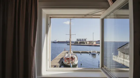 Et åbent vindu tilbyder en solrig udsigt over en havn med sejlbåde fortøjet ved en mole, et fjernt skib og bygninger på den fjerne kyst.