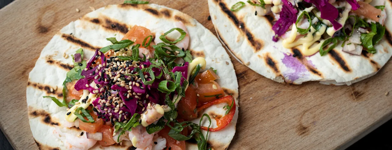 To grillede tortillas toppet med reker, tomater, strimlet rødkål, vårløk, sesamfrø og saus på et trebrett.