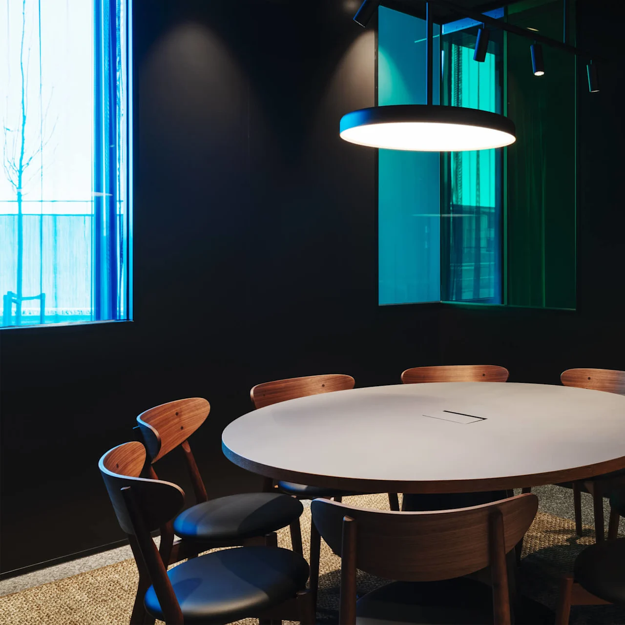 A round table surrounded by wooden chairs sits under a hanging light in a dimly lit room with a blue-tinted window.