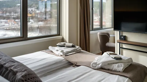 A neatly made bed with folded towels and robes rests near large windows revealing a scenic city view. Nearby, a flat-screen TV and a cushioned chair complete the modern hotel room.