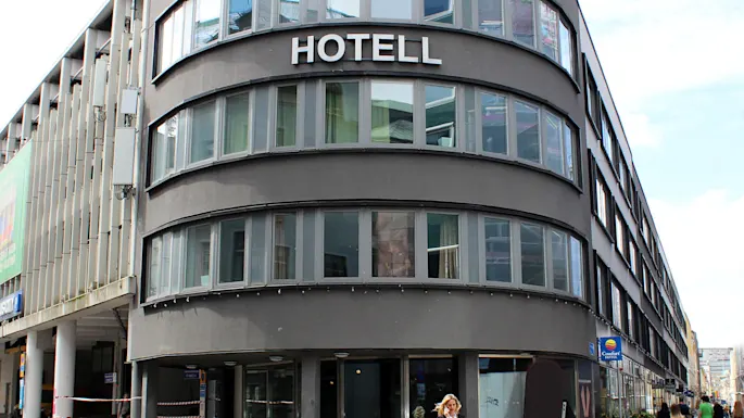 En buet moderne bygning mærket "HOTELL" med store vinduer; en person går på brostensbelagt gade udenfor. Omgivende bygninger og en klar himmel skaber en urban kontekst.