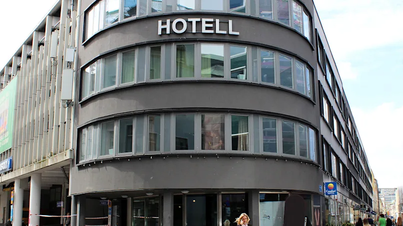 A curved modern building labeled "HOTELL" with large windows; a person walks on the cobblestone street outside. Surrounding buildings and a clear sky provide an urban setting.