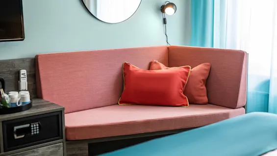 Corner sofa with red cushions sits beside a coffee table in a room. Lamp is mounted above, and a mirror hangs on light blue wall near window.