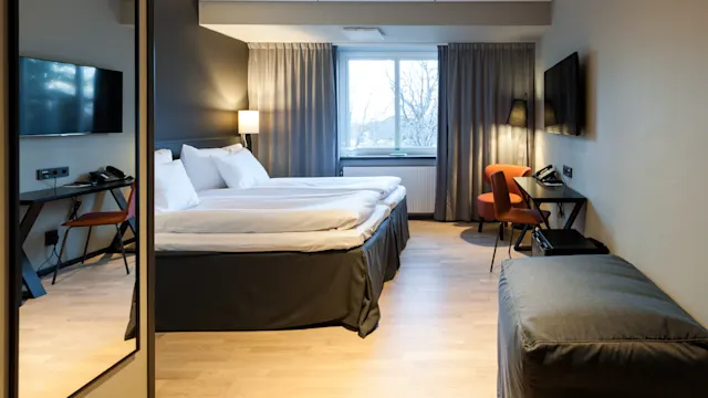 Siisti sänky sijaitsee modernissa hotellihuoneessa, jossa on puulattia, kaksi taulutelevisiota ja työpöytä oranssilla tuolilla. Suuri ikkuna päästää sisään luonnonvaloa.