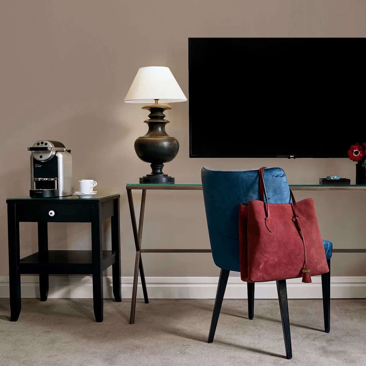 A blue chair with a red bag rests by a glass-topped desk, featuring a lamp, red flowers, and books. Nearby, a small table holds a coffee maker and cup under a wall-mounted TV.