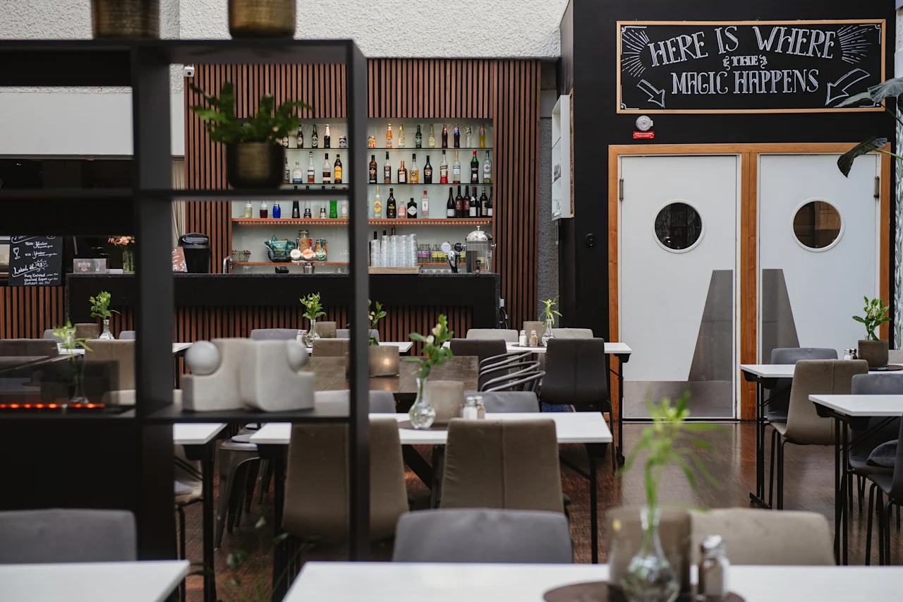 En restauranginteriör har bord med stolar, växter och en bakvägg med hyllor av flaskor. Ovanför en dörr står en skylt med texten "HERE IS WHERE THE MAGIC HAPPENS."
