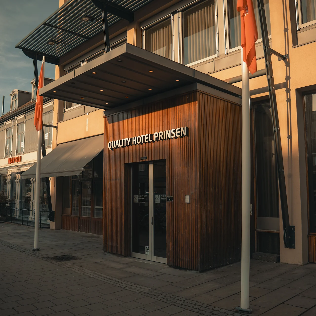Hotellin sisäänkäynti, jossa lukee "QUALITY HOTEL PRINSEN" puuverhoilulla, sijaitsee rauhallisella kadulla, jossa on toinen rakennus merkinnällä "NAPOLI" lähistöllä.