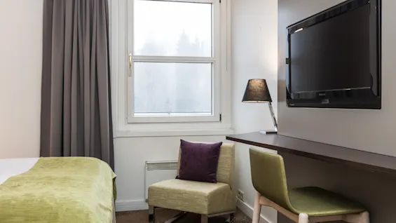 A wall-mounted TV is off next to a desk with a green chair, lamp, and window. A bed with a green blanket and a cushioned chair with a purple pillow are nearby.