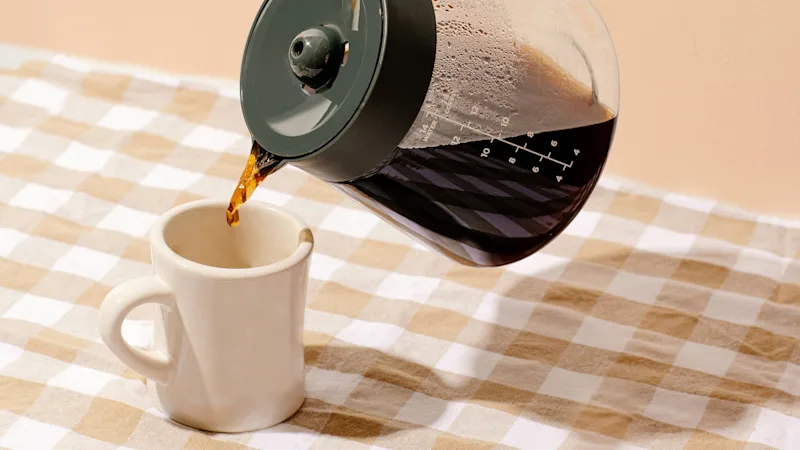 En hånd heller kaffe fra en glasskanne i en hvit kopp på en rutete duk, mot en ferskenfarget vegg.