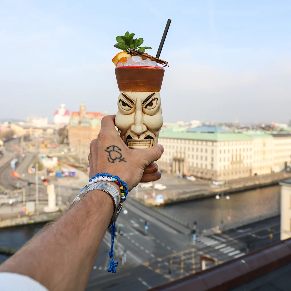 A hand holds a tiki cocktail mug with a garnished drink, overlooking an urban landscape from a rooftop.
