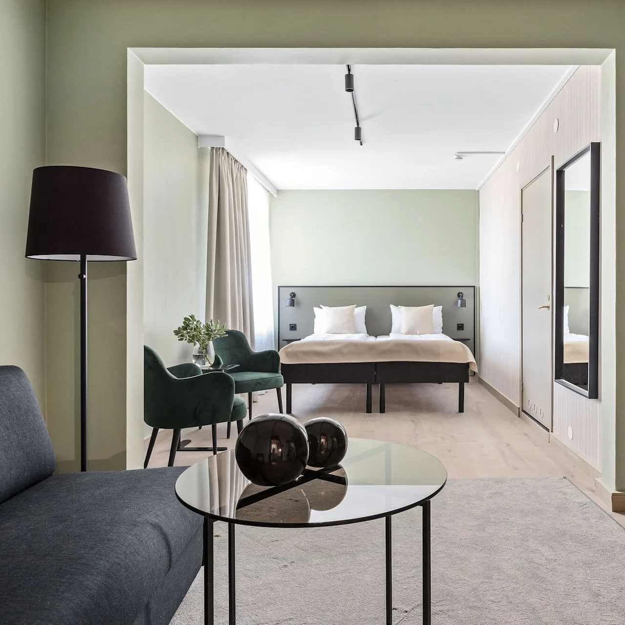 Ett modernt, minimalistiskt sovrum med dubbelsäng, gröna väggar och naturligt ljus från stora fönster, samt en mörk soffa, glasbord och elegant golvlampa.