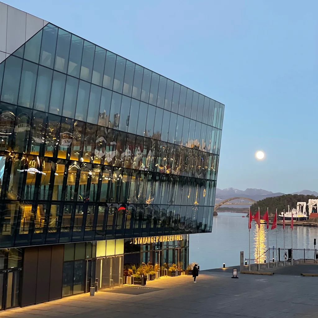 Moderni lasirakennus, STAVANGER KONSERTHUS, veden äärellä täydenkuun aikaan hämärässä. Henkilö kävelee lähellä.