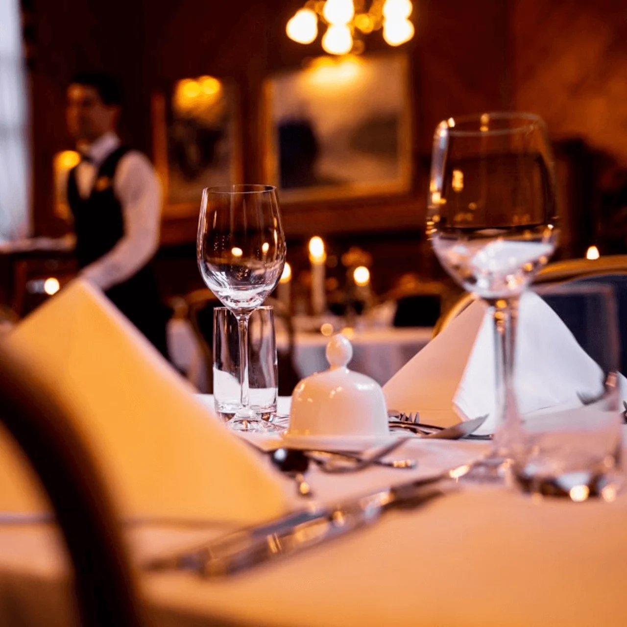 Dining table is set with wine glasses and cutlery, lit by candles in an elegant restaurant. A waiter stands in the background, enhancing the sophisticated ambiance.