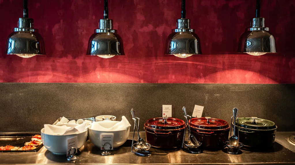 Boller og gryder er arrangeret på en bordplade, med suppeskeer hvilende i nærheden. Overhead oplyser fire metalliske pendellamper scenen mod en varm, rød baggrund.