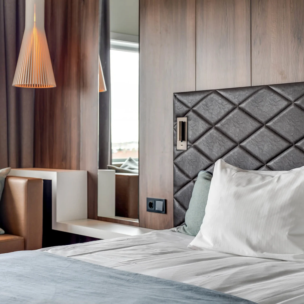 A modern bedroom with a neatly made bed, diamond-patterned headboard, wood paneling, a pendant lamp, and a long curtain. Contemporary, cozy atmosphere.