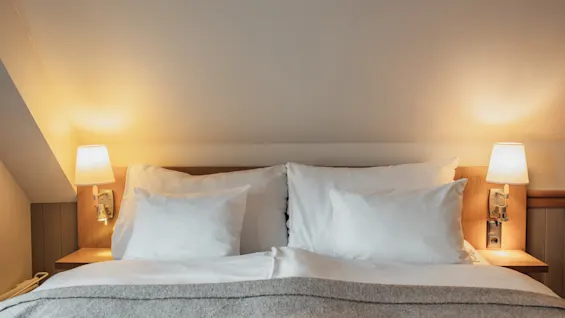 A cozy bed with white linens and a grey blanket, illuminated by warm bedside lamps in a room with a sloped ceiling.