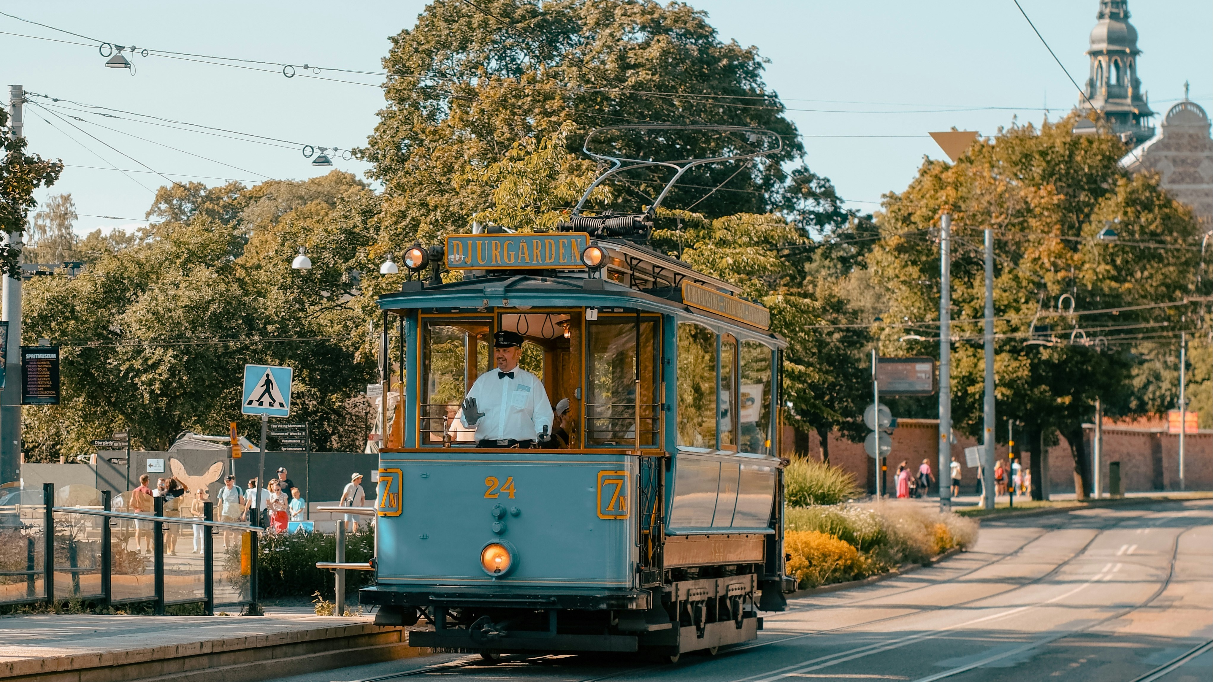 A light blue tram with a driver in uniform travels down a street on a sunny day. DJURGARDEN is visible on the tram.