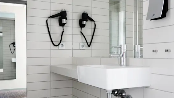 Dual wall-mounted hairdryers are secured above a white tiled bathroom sink, with mirrors reflecting the scene. Metal fixtures, outlets, and minimalistic design enhance the clean, modern aesthetic.