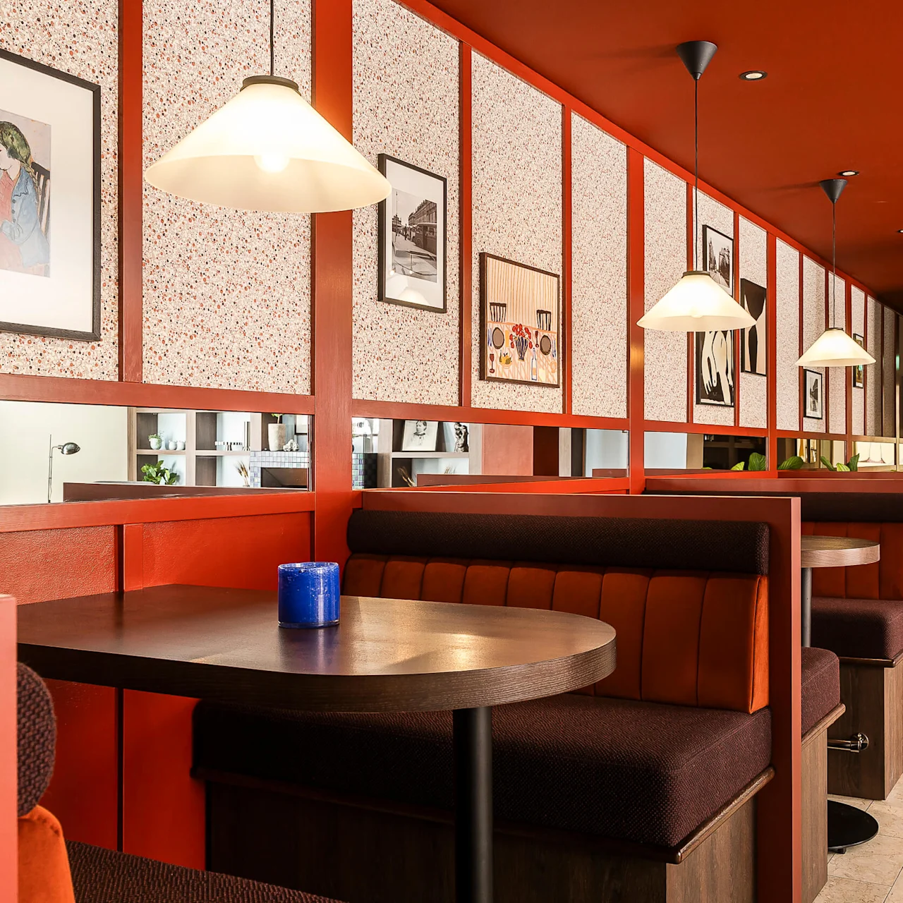 Tomma restaurangbås med belysning ovanför. Inramade konstverk hänger på väggen. Stämningen är varm med en modern inredning.