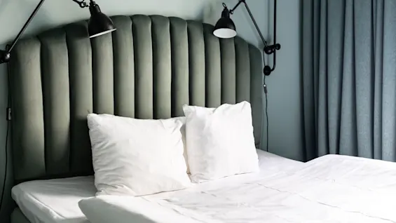 En koselig hotellseng med en grønn sengegavl, hvite puter og to leselamper, som inviterer til en god natts søvn.