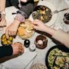 Several hands reach for food on a table. Olives, bread, and dishes including a green and black dish are present. Wine glasses and a lit candle add to the dining atmosphere.